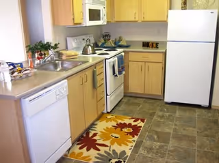 Small kitchen with light wood cabinets, white appliances (refrigerator, stove, microwave, dishwasher), sink and a floral floor rug.