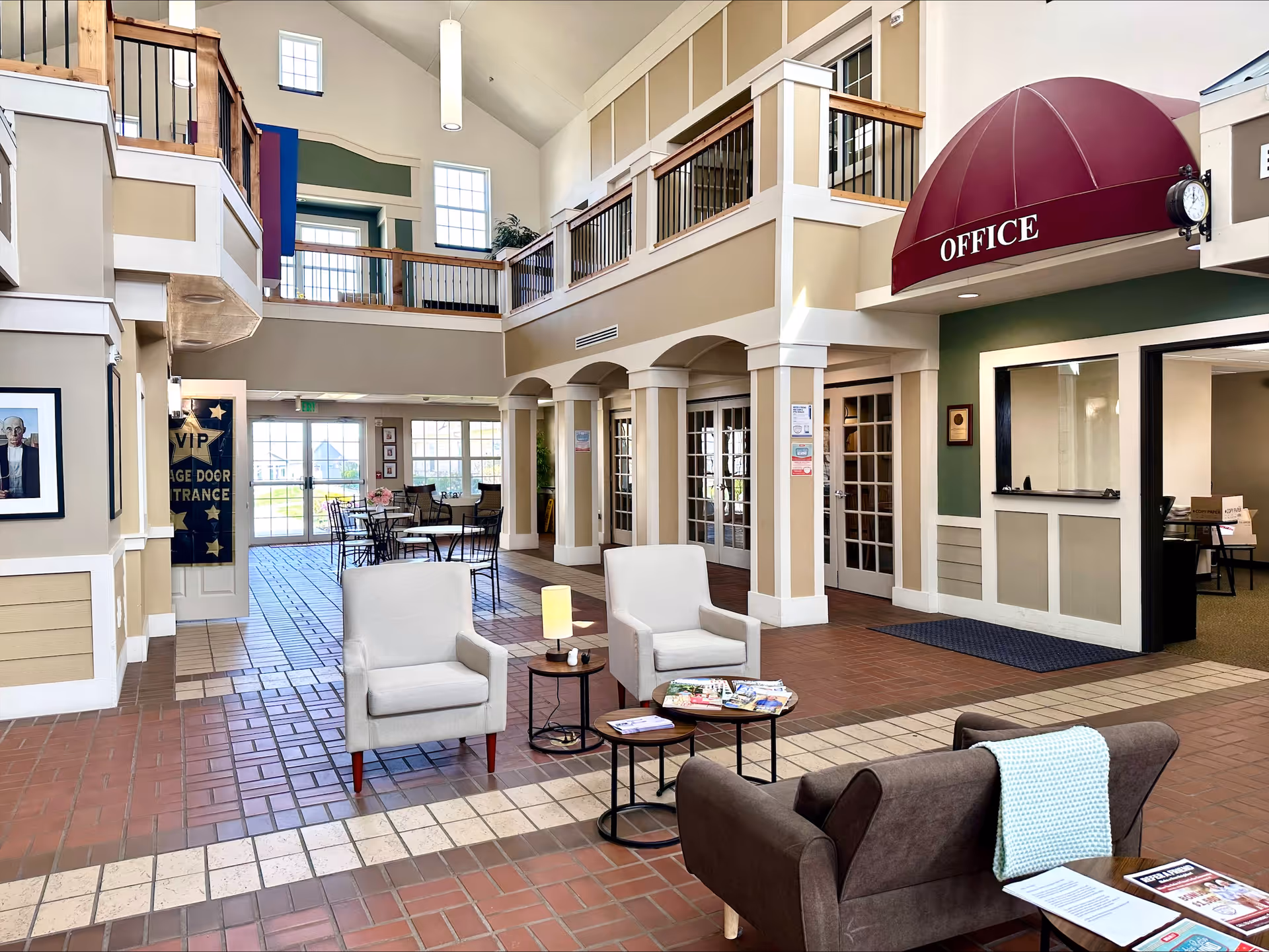 Spacious senior living lobby with seating, tables, a reception office under a red awning, and a second-floor balcony.