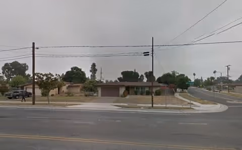 Street view of a single-story residential building with a garage, surrounded by trees and bushes, under an overcast sky.