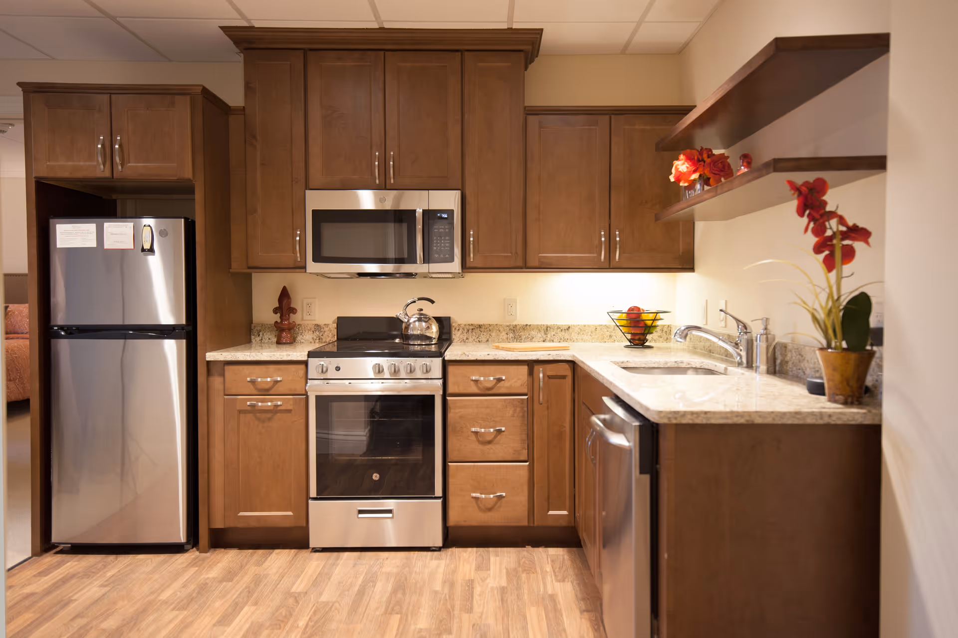 A modern kitchen with wooden cabinets, stainless steel refrigerator, oven with stove, microwave, and dishwasher. The countertop is granite with a sink and faucet. There are two wooden shelves on the right wall with decorative red flowers and a fruit basket on the counter. The floor is wood, and the kitchen is well-lit with under-cabinet lighting.