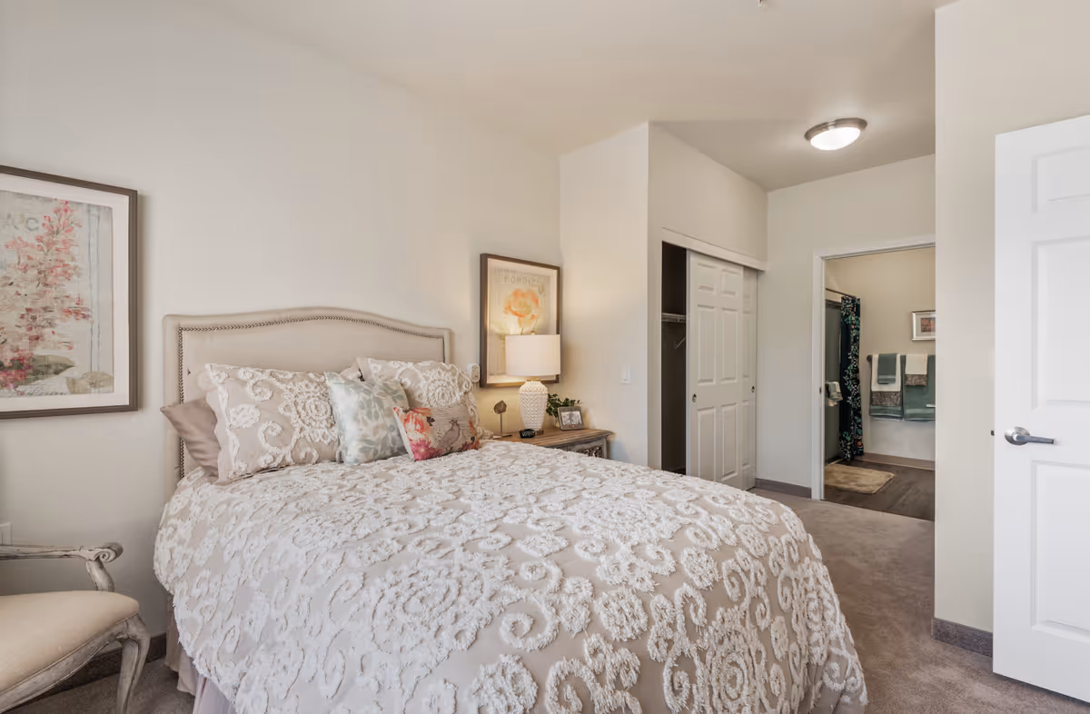 A cozy bedroom with a beige upholstered bed adorned with decorative pillows and a textured bedspread. There is a wooden nightstand with a lamp, a framed picture on the wall, and a chair with a cushioned seat. The room has light-colored walls and carpeted floor. A closet with sliding doors and an open door leading to a bathroom with towels and a shower curtain are visible.
