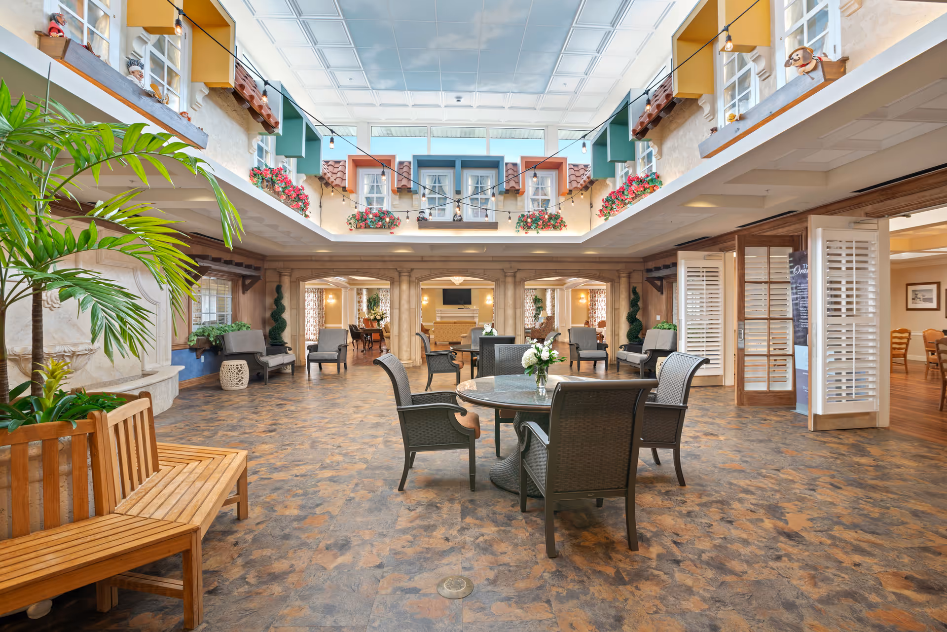 Spacious indoor common area with a high ceiling featuring a sky and clouds mural. The room has a round table with four chairs in the center, wooden benches, potted plants, and multiple seating areas with armchairs. The walls are decorated with window-like structures and flower boxes, and there are string lights hanging across the ceiling. The area has an inviting and bright atmosphere.