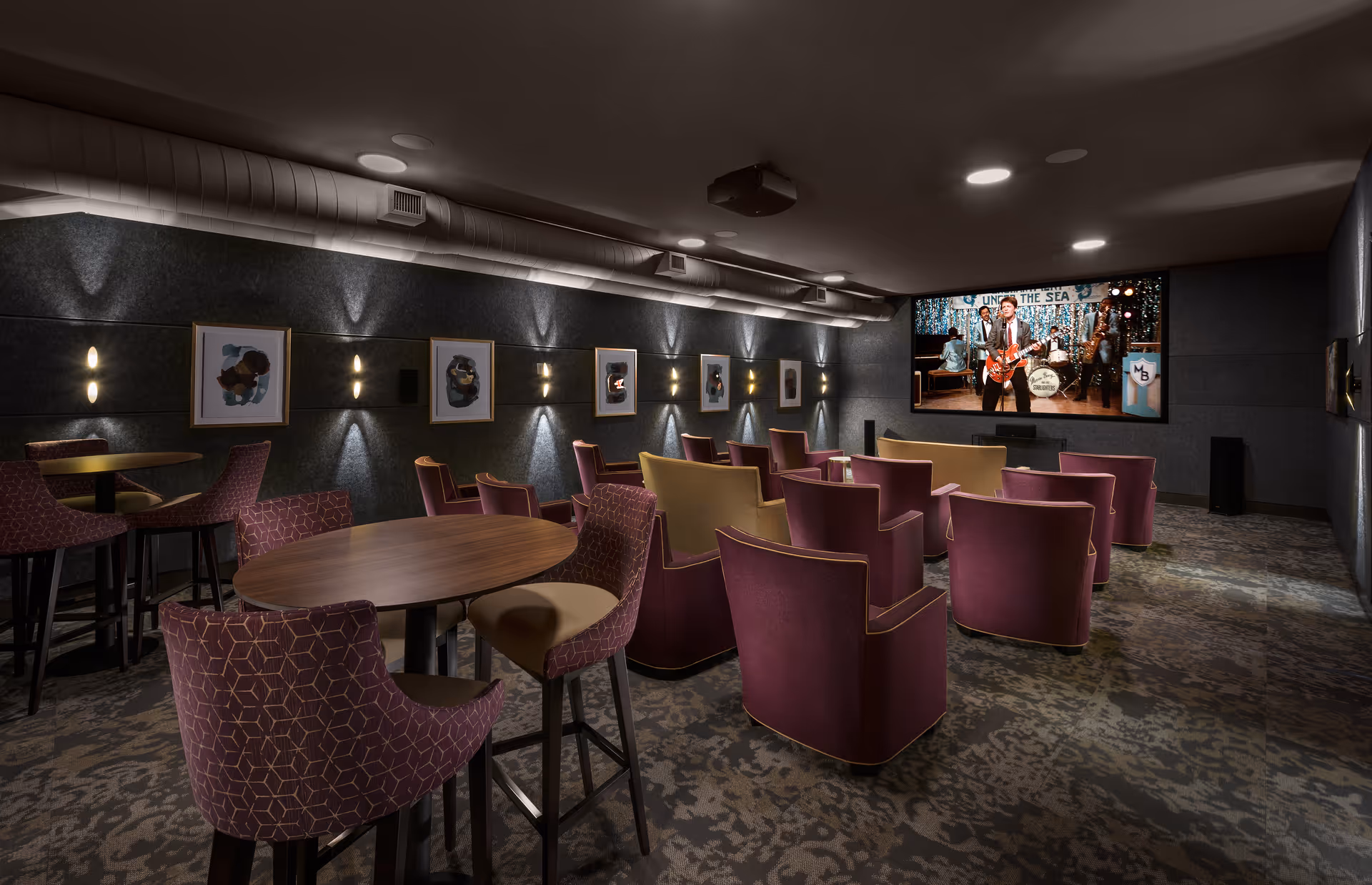 A cozy movie theater room with multiple rows of comfortable purple and beige chairs facing a large screen showing a band performing. The walls are decorated with framed abstract art and soft wall lights, and there are a few round tables with chairs in the foreground.