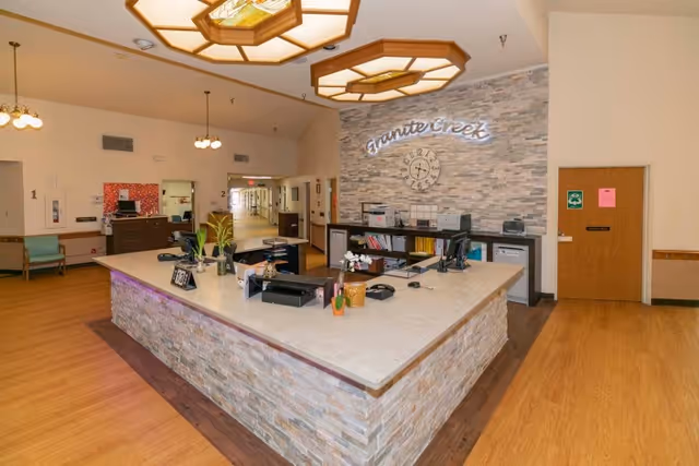 Reception area inside Granite Creek Health and Rehabilitation facility with a large stone-faced desk, office supplies, plants, and a wall clock. The wall behind the desk features the facility name 'Granite Creek' in illuminated letters. The area has wooden flooring, ceiling lights, and a hallway leading further into the building.