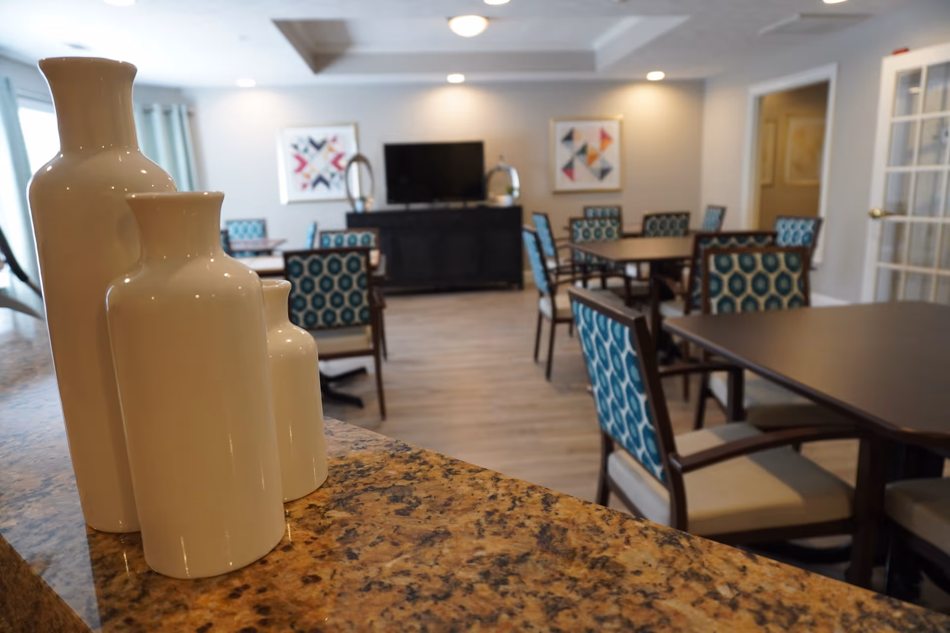 Interior view of a common area in a senior living facility with multiple tables and chairs featuring blue patterned upholstery. In the foreground, three white decorative vases are placed on a granite countertop. The room has light-colored walls, framed abstract artwork, and a television on a black cabinet against the far wall.