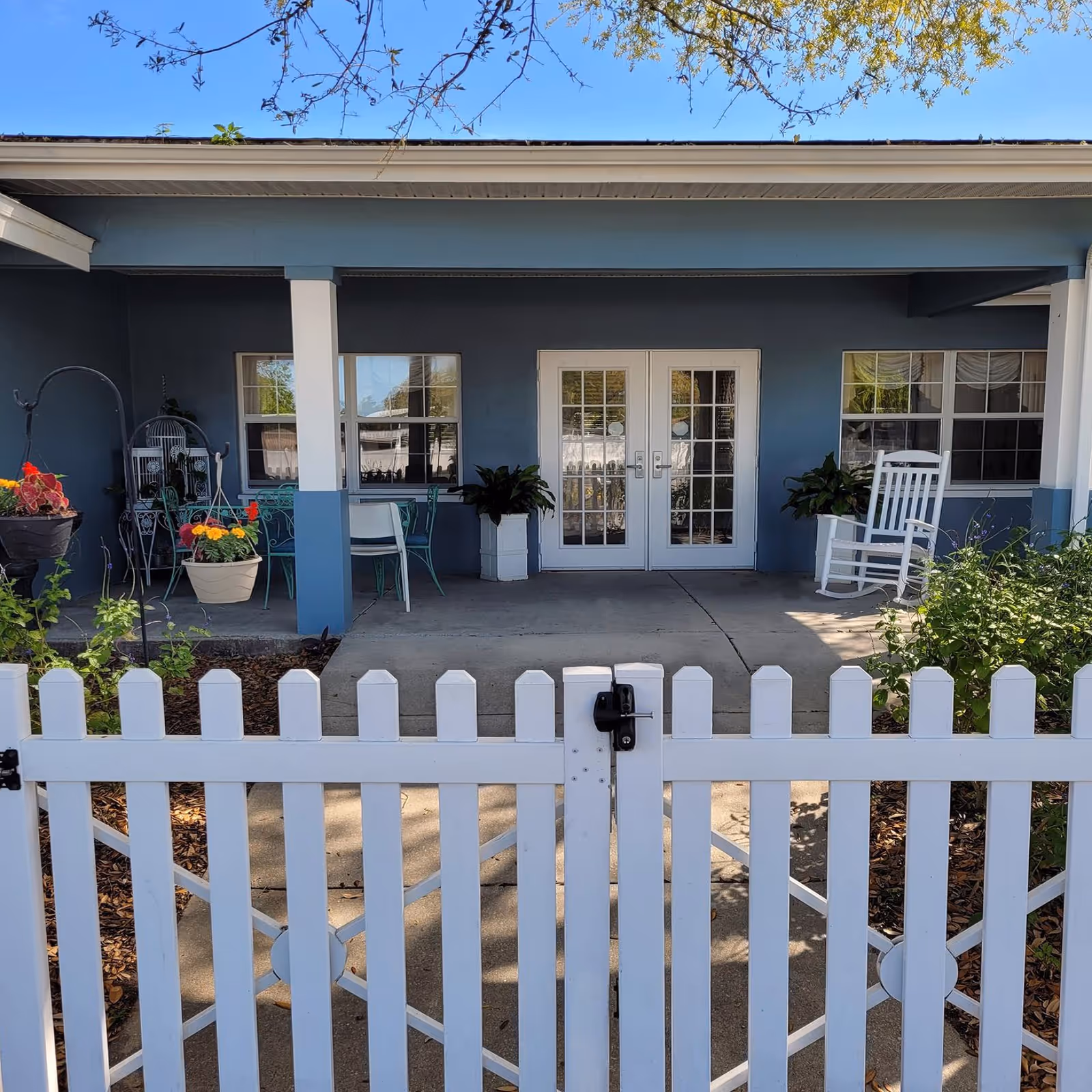 A front porch area of a building with a white picket fence gate in the foreground. The porch has two white columns, a pair of white French doors, two windows, a white rocking chair, potted plants, and a small table with chairs. The building exterior is painted blue with white trim.
