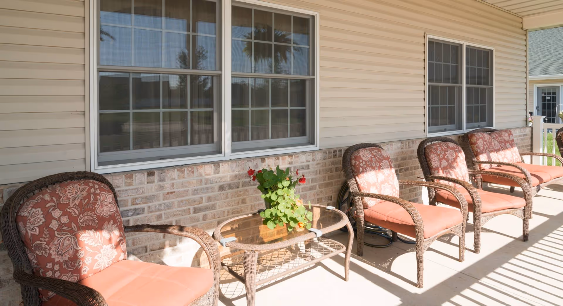 A sunlit outdoor patio area with four wicker chairs featuring floral patterned cushions and a small glass-top wicker table with a potted plant. The patio is adjacent to a building with beige siding and brick lower walls, and has two large windows.