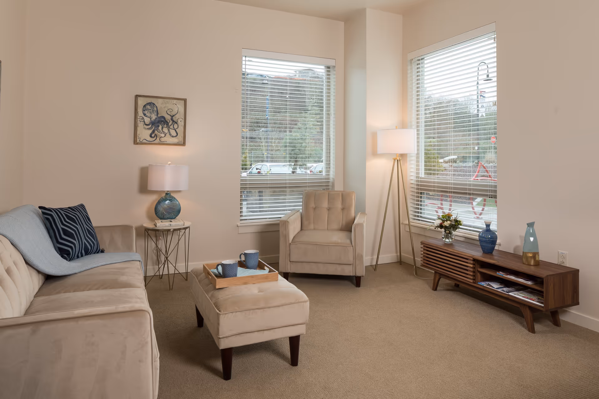 A cozy living room with beige walls and carpet. The room features a beige sofa with a blue pillow and throw, a matching beige armchair, and a beige ottoman with a wooden tray holding two blue mugs. There is a small round side table with a lamp next to the sofa, and a tall floor lamp near the corner. A wooden media console with decorative vases and flowers sits under two large windows with white blinds, through which an outdoor parking area and greenery are visible. A framed octopus artwork hangs on the wall above the side table.