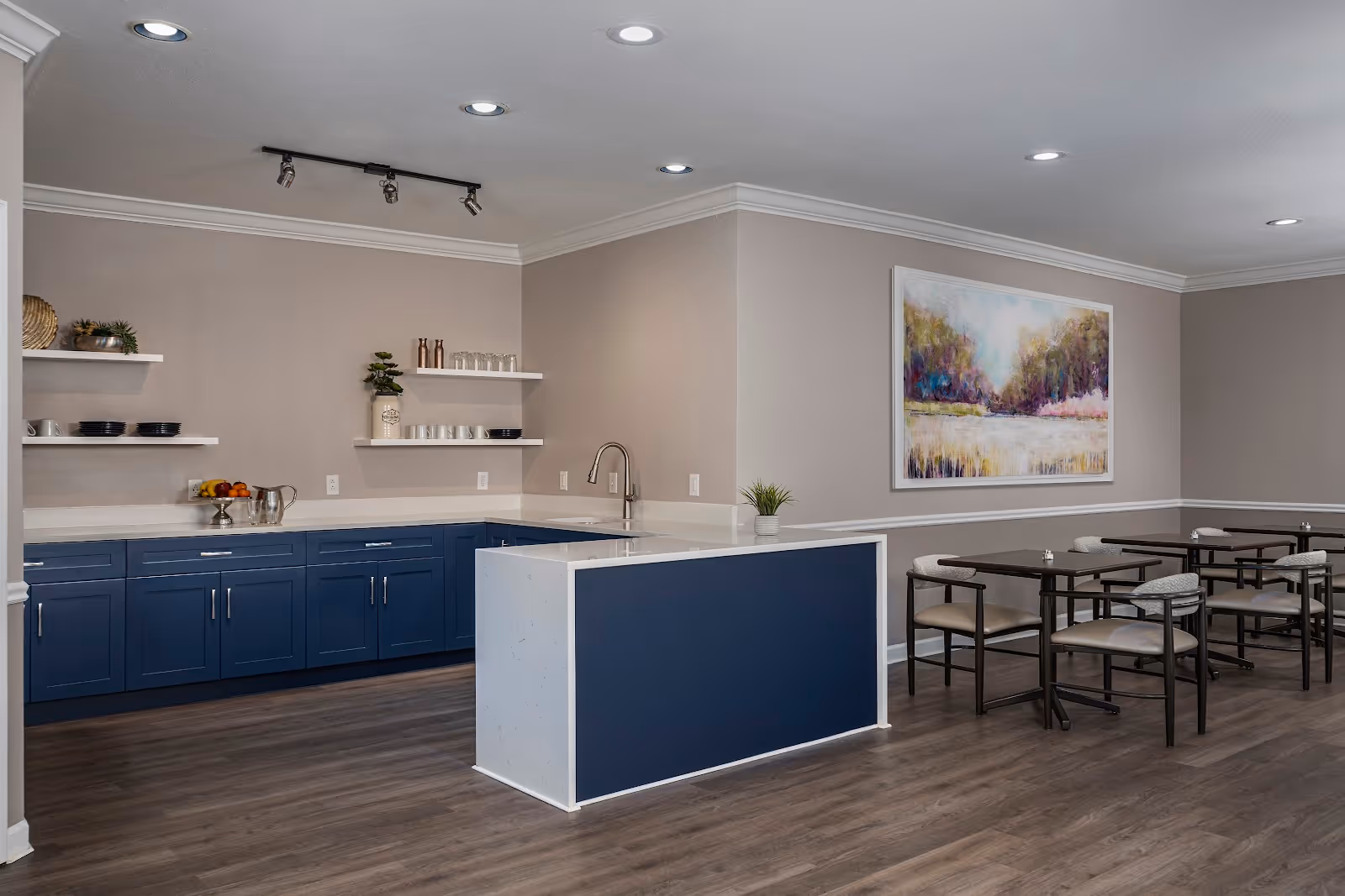A modern assisted living facility dining area with a kitchen counter featuring blue cabinets and white countertops. The kitchen has open shelves with dishes and decorative items. Adjacent to the kitchen are several dark wood dining tables with chairs, and a large colorful landscape painting on the wall. The room has recessed lighting and wood flooring.