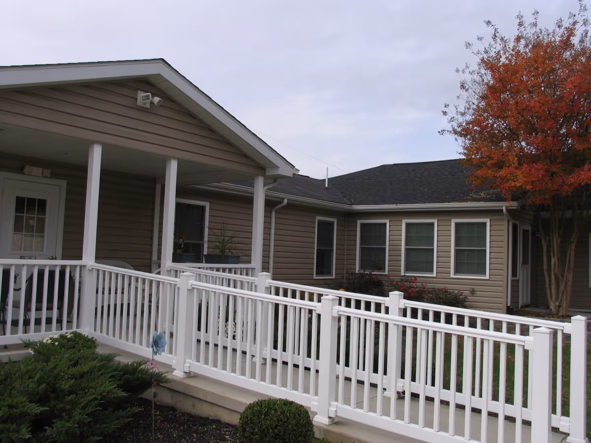 Single-story assisted living building exterior with a covered entrance, white accessibility ramp railing, windows, and a tree with red leaves.