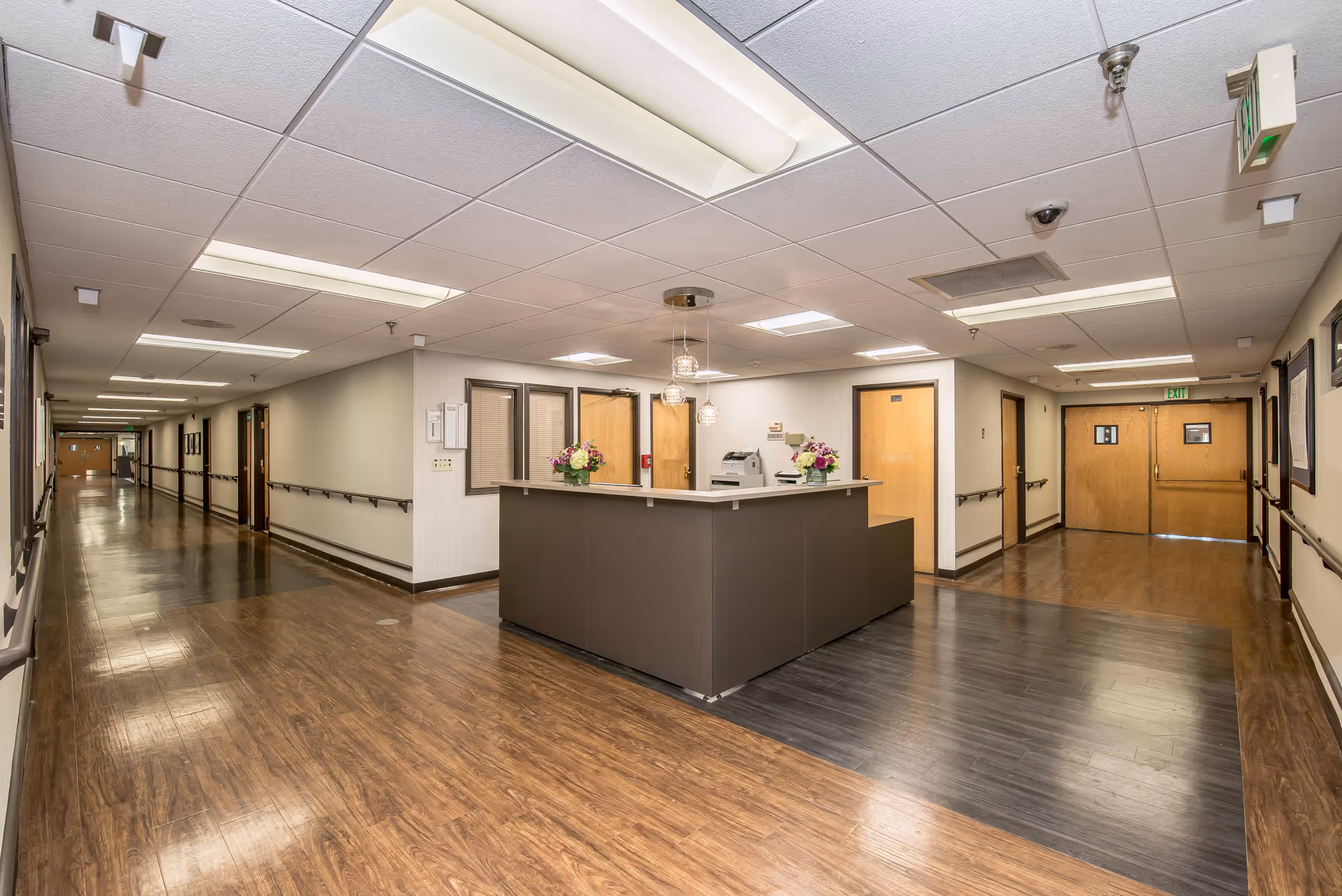 A clean and well-lit hallway in a senior living facility with wood flooring and beige walls. In the center is a reception desk decorated with flower arrangements. Multiple doors and handrails line the hallway, and fluorescent ceiling lights illuminate the space.