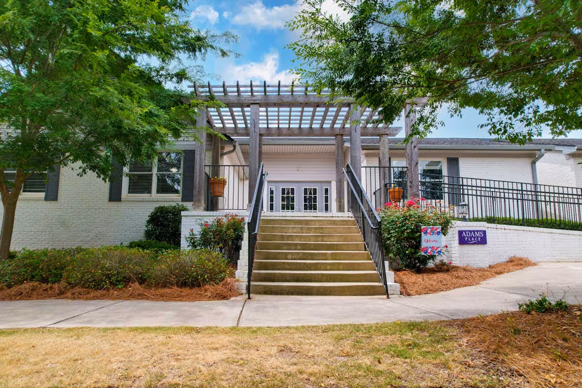 Front entrance of a white brick building with stairs leading up to double glass doors under a wooden pergola. There are bushes and trees on either side of the stairs, and a sign on the right side reads 'ADAMS PLACE'.