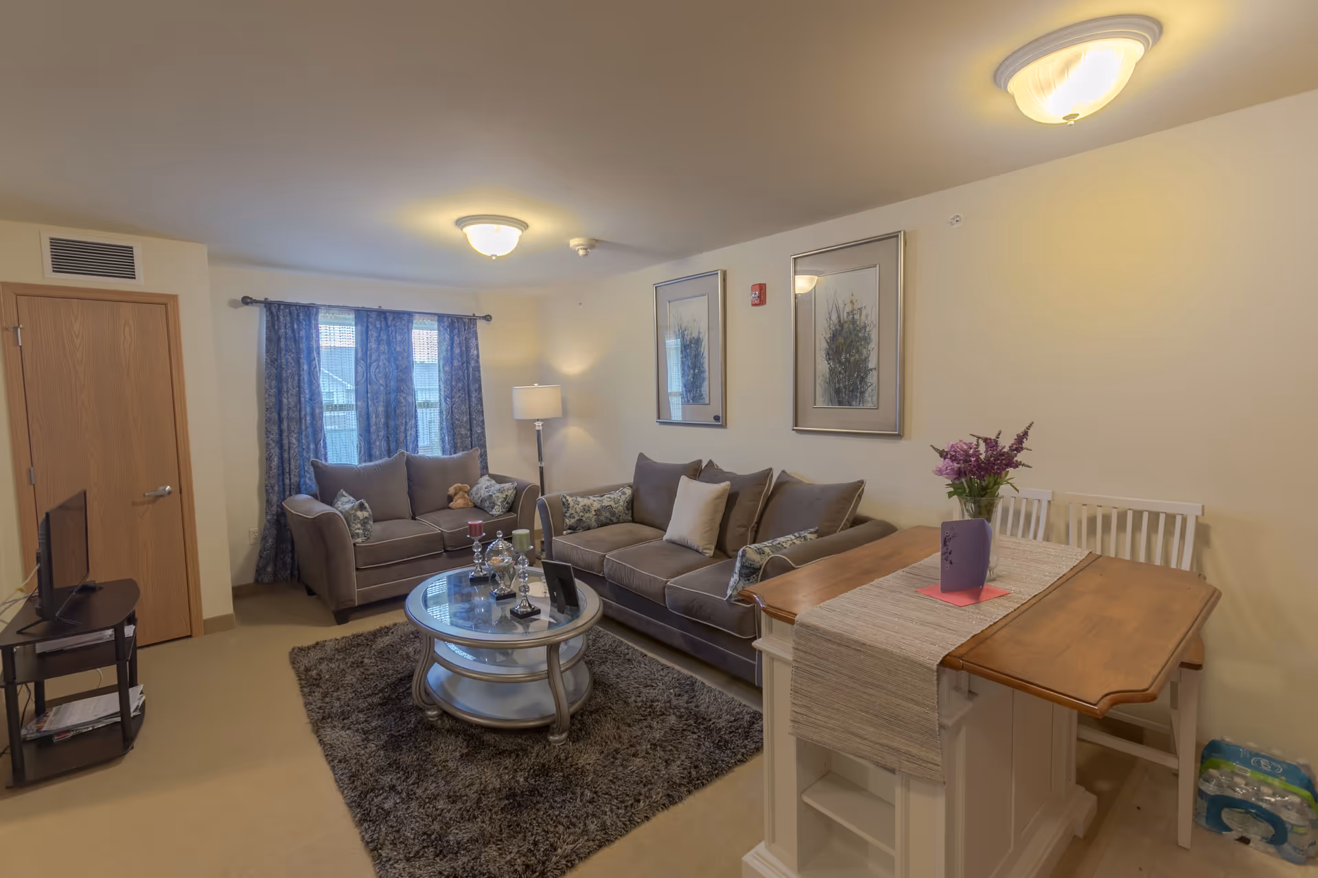 Cozy living room featuring two grey sofas, a round glass coffee table on a shag rug, and a small dining table with chairs.