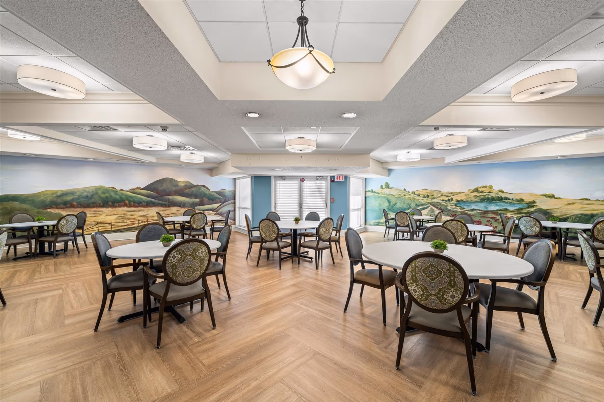 Spacious dining room with round tables and patterned chairs, wood-look flooring and a panoramic landscape mural on the walls.