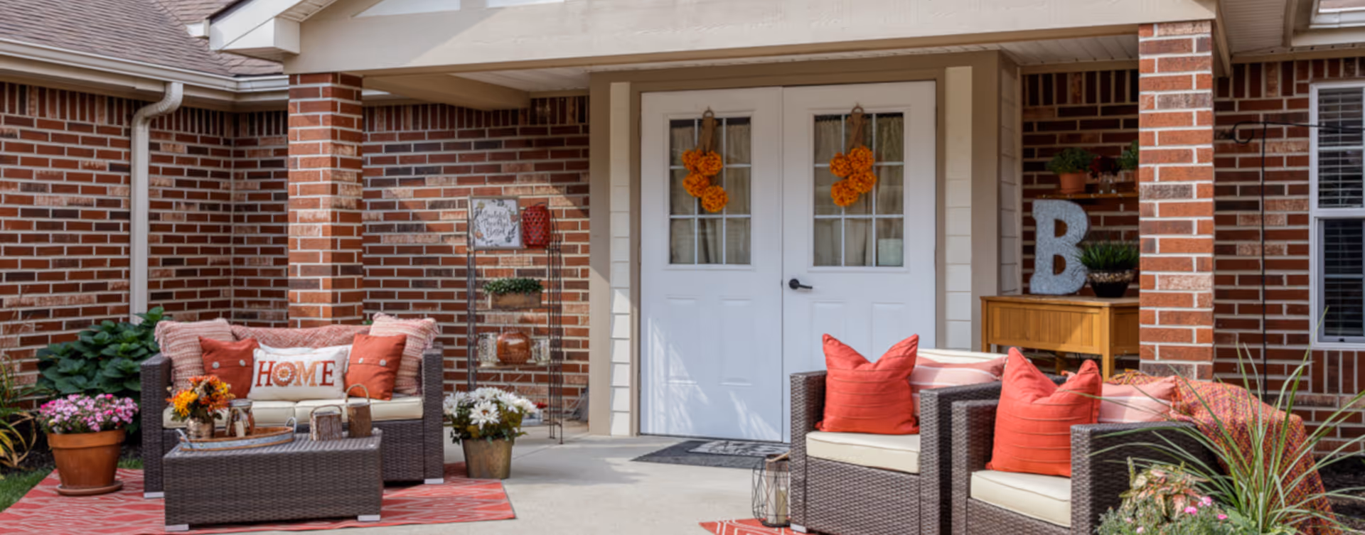 Outdoor patio area at Bickford of West Des Moines featuring wicker furniture with red and beige cushions, decorative pillows including one with the word 'HOME', potted plants, flowers, and a brick exterior wall with white double doors adorned with orange wreaths.
