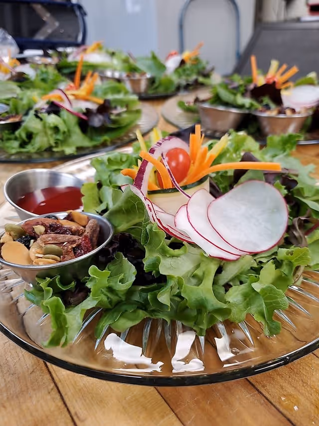 Close-up view of fresh salad plates arranged on a wooden table, each plate containing leafy greens, sliced radishes, shredded carrots, cherry tomatoes, a small cup of mixed nuts and seeds, and a small cup of red dressing.