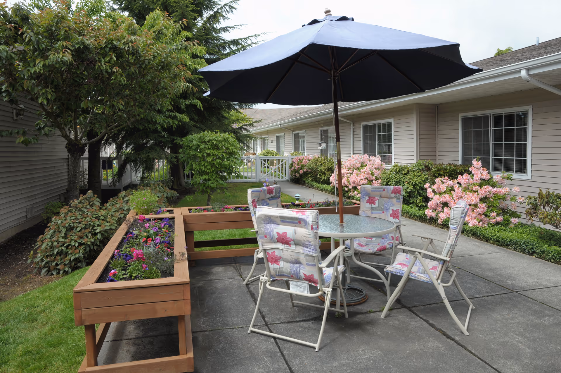Outdoor courtyard patio with a round table, umbrella and cushioned chairs beside raised flower planters and the facility building.