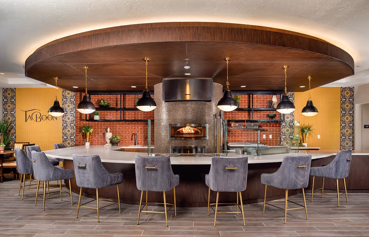 Modern kitchen area with a curved white countertop surrounded by gray upholstered bar stools with gold legs. Above the counter, there are six black pendant lights hanging from a large wooden ceiling fixture. In the center, a built-in pizza oven with a fire burning inside is visible, set against a tiled wall with red and yellow tiles and decorative patterned accents. Potted plants and decorative items are placed on shelves and counters.