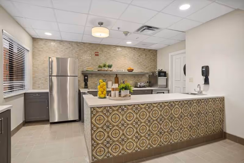 A modern kitchen area with a stainless steel refrigerator, gray cabinets, a tiled backsplash, and a patterned tile front on the kitchen island. The island countertop has decorative items including a jar of lemons and a tray with plants and cups. The room has a window with blinds, ceiling lights, and a wall-mounted soap dispenser near a small sink.