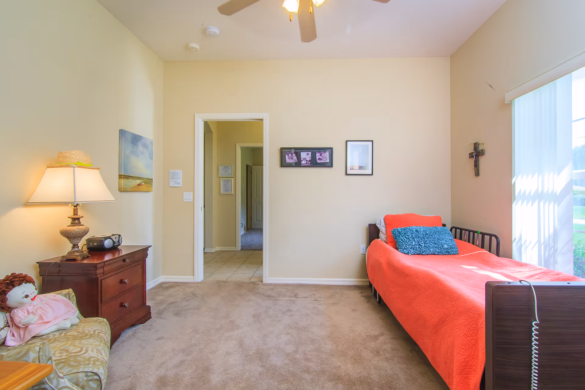 A bright bedroom with beige walls and carpeted floor. The room features a single bed with a red bedspread and a blue pillow, positioned next to a large window with vertical blinds. On the right wall above the bed, there is a cross hanging. On the left side of the room, there is a wooden nightstand with a lamp and a small radio, next to an armchair with a doll sitting on it. The room has a ceiling fan with lights and an open doorway leading to a tiled hallway.