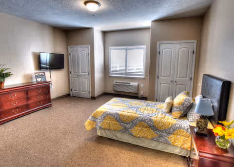 A spacious furnished bedroom with a bed covered in yellow patterned bedding, a nightstand and lamp, a dresser with a wall-mounted TV, a window, and two doors.