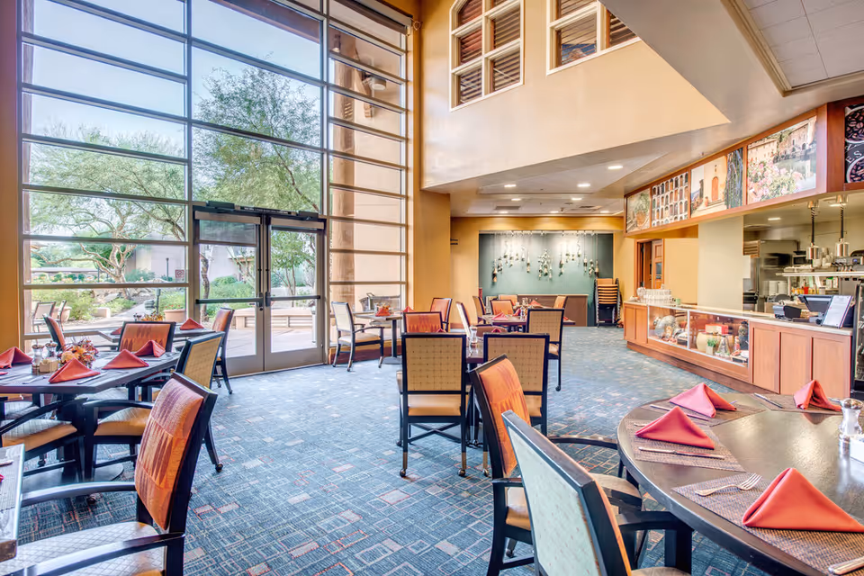 Bright dining room with large floor-to-ceiling windows showing outdoor greenery. Several tables are set with red folded napkins and chairs arranged around them. The room has a carpeted floor and a serving counter on the right side with kitchen equipment visible behind it.
