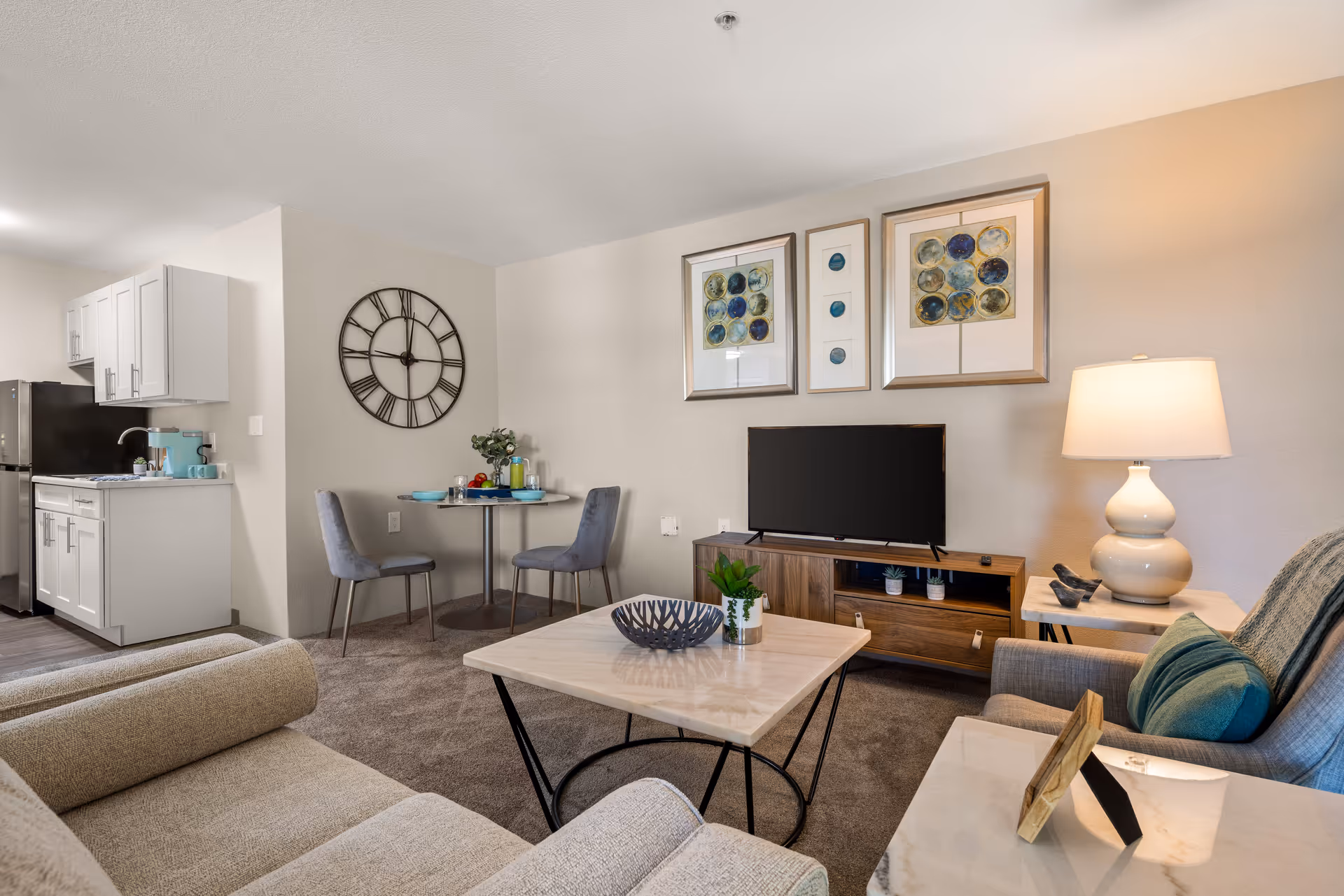 A cozy living room and kitchen area in a senior living community apartment. The living room features a beige sofa, a coffee table with decorative items, a TV on a wooden stand, and a side table with a lamp and photo frame. On the wall above the TV are three framed abstract artworks. Adjacent to the living room is a small dining area with a round table and two gray upholstered chairs. The kitchen has white cabinets, a stainless steel refrigerator, and a countertop with a coffee maker and cups. A large decorative wall clock is mounted on the wall near the dining area.