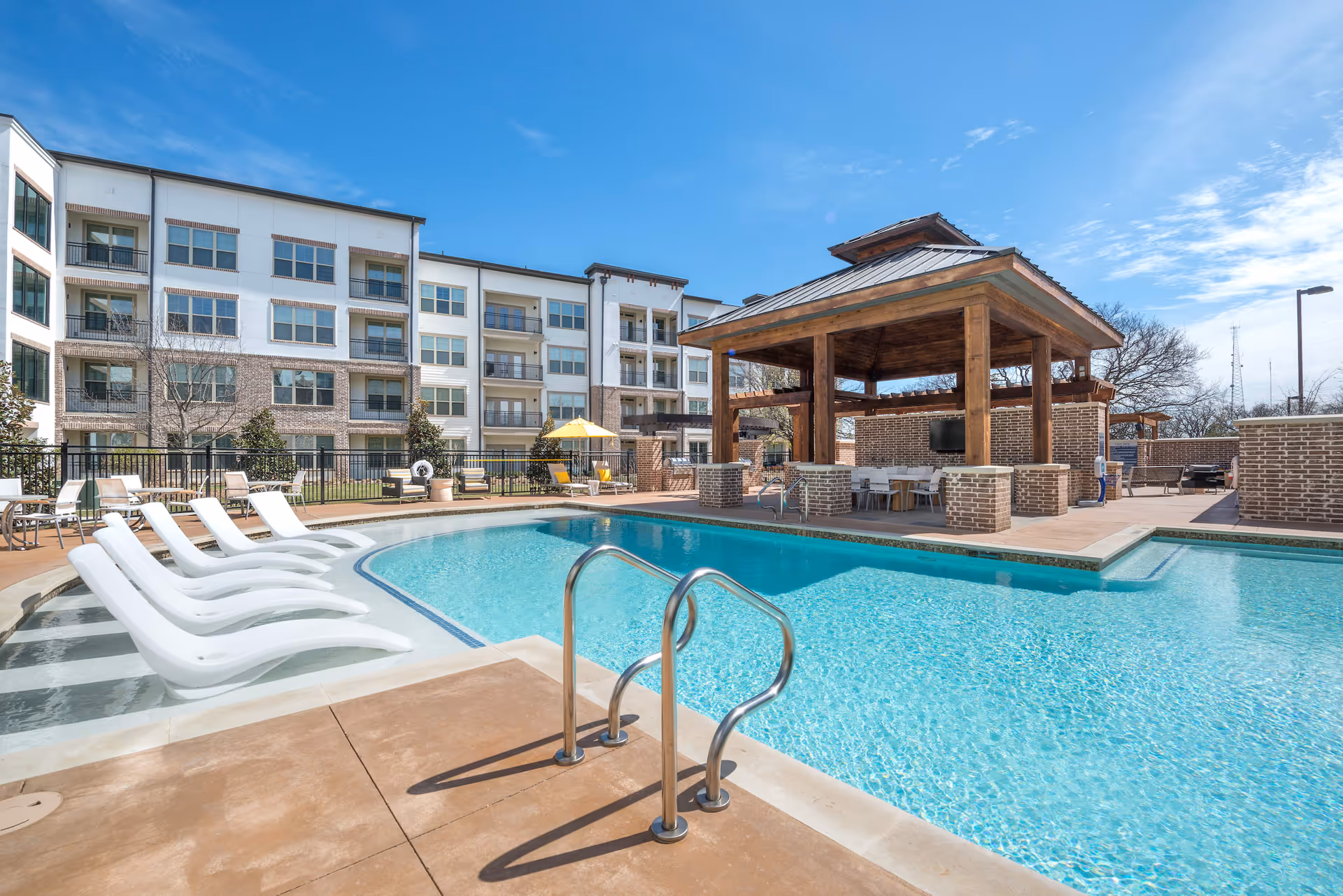 Outdoor swimming pool area with white lounge chairs partially submerged in the water, a wooden gazebo with seating and a TV, and a multi-story residential building in the background under a clear blue sky.