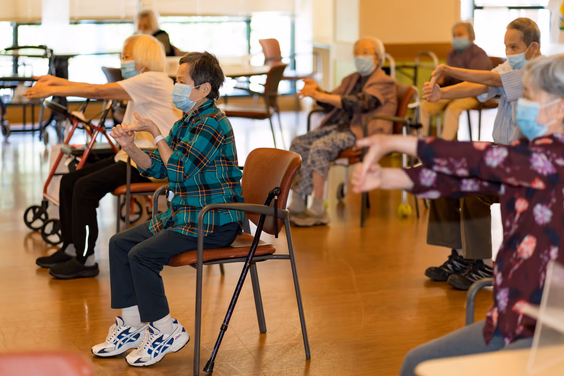 A group of elderly people wearing face masks sitting on chairs in a spacious room, participating in a seated exercise session with arms extended forward.