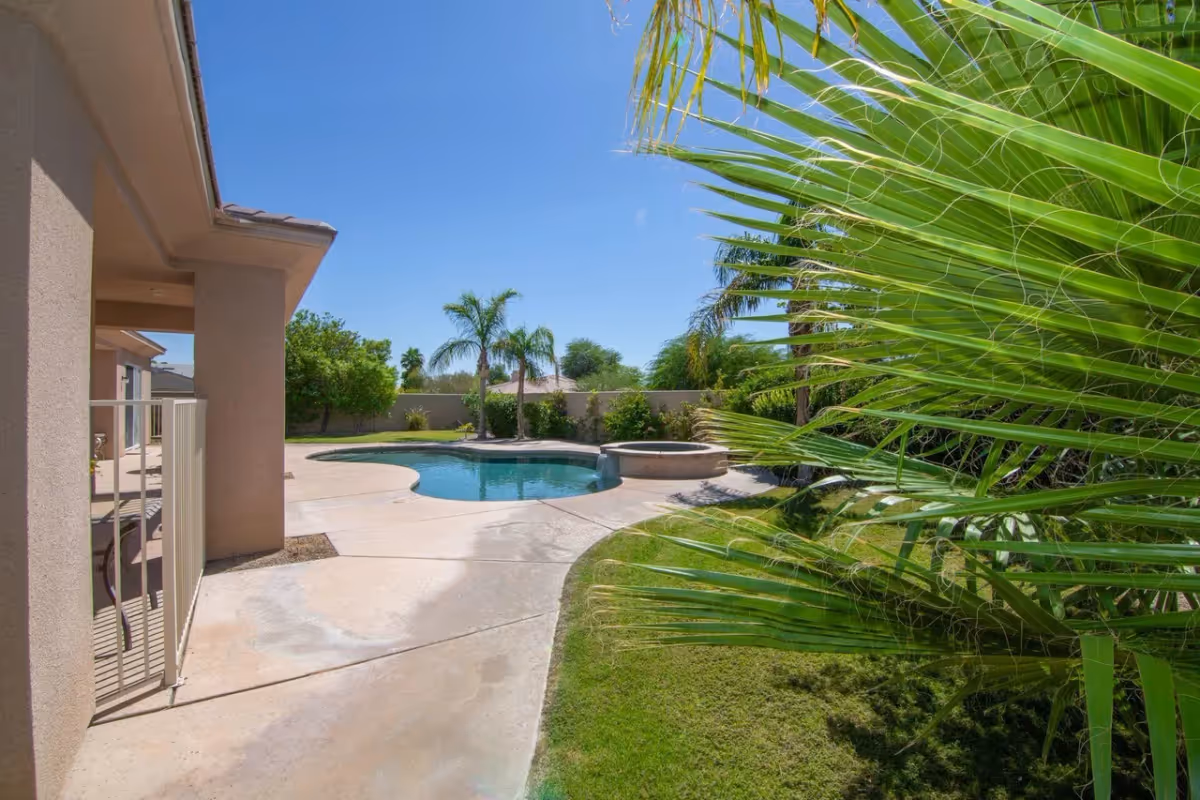 Outdoor view of a sunny backyard area with a swimming pool and a hot tub surrounded by palm trees and green grass, adjacent to a beige building with a covered patio.