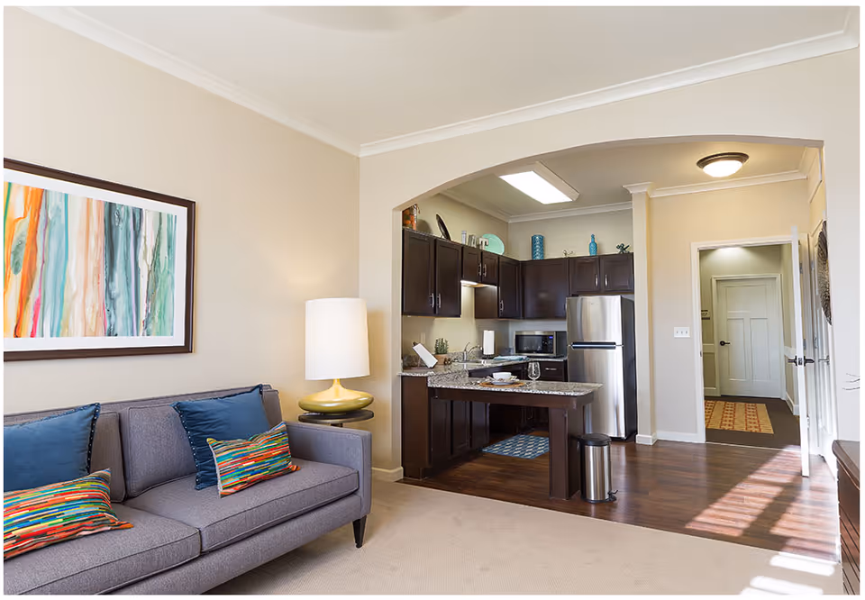A cozy living room area with a gray sofa adorned with colorful pillows and a modern lamp on a side table. Adjacent to the living room is an open kitchen with dark wood cabinets, granite countertops, stainless steel refrigerator, microwave, and a small island with a place setting. The space features light beige walls, hardwood flooring in the kitchen area, and carpet in the living room. A hallway with doors is visible in the background.