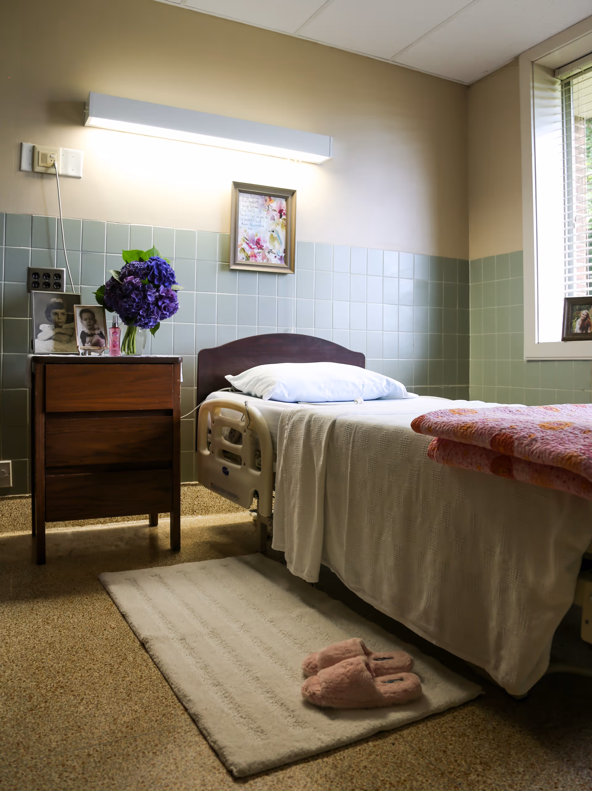 A single hospital-style bed with white bedding and a pink quilt folded at the foot, placed next to a wooden nightstand with framed photos and a vase of purple flowers. A white rug with pink slippers is on the floor beside the bed. The room has green tiled walls halfway up and a window with blinds letting in natural light.