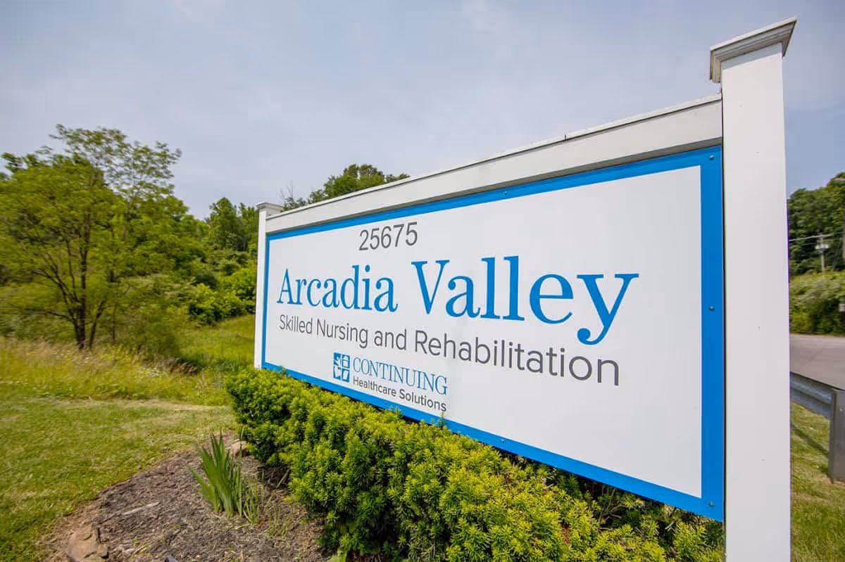 Outdoor view of a large white and blue sign for Arcadia Valley Skilled Nursing and Rehabilitation, surrounded by greenery and trees under a partly cloudy sky.