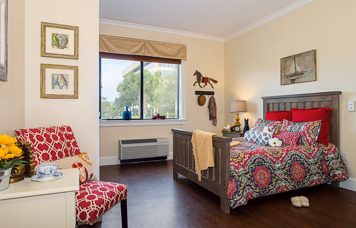 A cozy bedroom in a senior living facility featuring a wooden bed with a colorful patterned bedspread and red pillows. There is a bedside table with a lamp and decorative items, a window with a view of trees and a building outside, and a red patterned chair next to a small white table with a teacup and flowers. The walls are decorated with framed bird pictures and a wooden horse wall hanging.