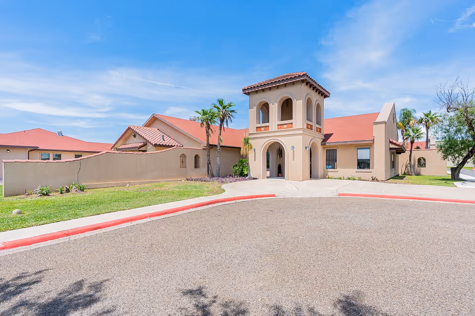 Front exterior of a beige stucco assisted living building with a red tile roof, arched two-story tower, palm trees, and a circular driveway.