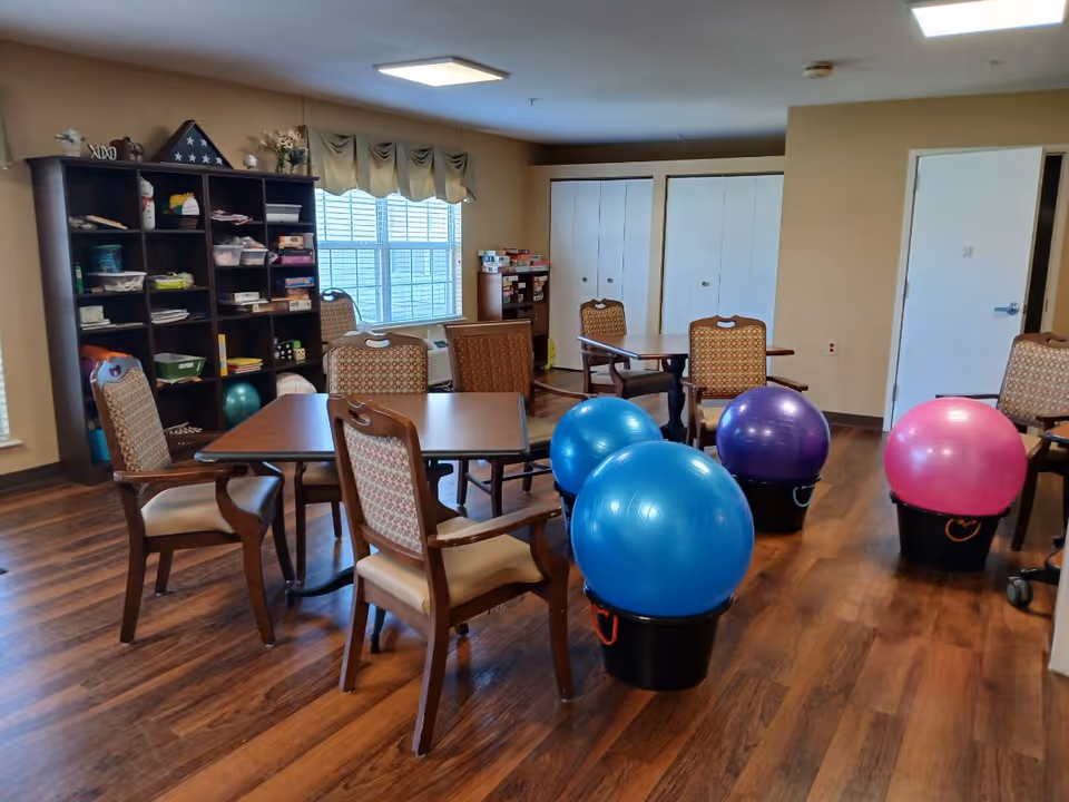 A senior living common room with tables and chairs, shelving, and several large colorful exercise balls on the floor.