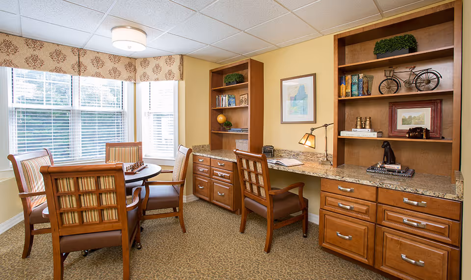 A cozy room with a round table and four wooden chairs with striped cushions near a large window with blinds and a patterned valance. On the right side, there is a built-in wooden desk with granite countertop, two chairs, and shelves holding books, decorative items, and framed pictures. The walls are painted light yellow and the floor is carpeted.