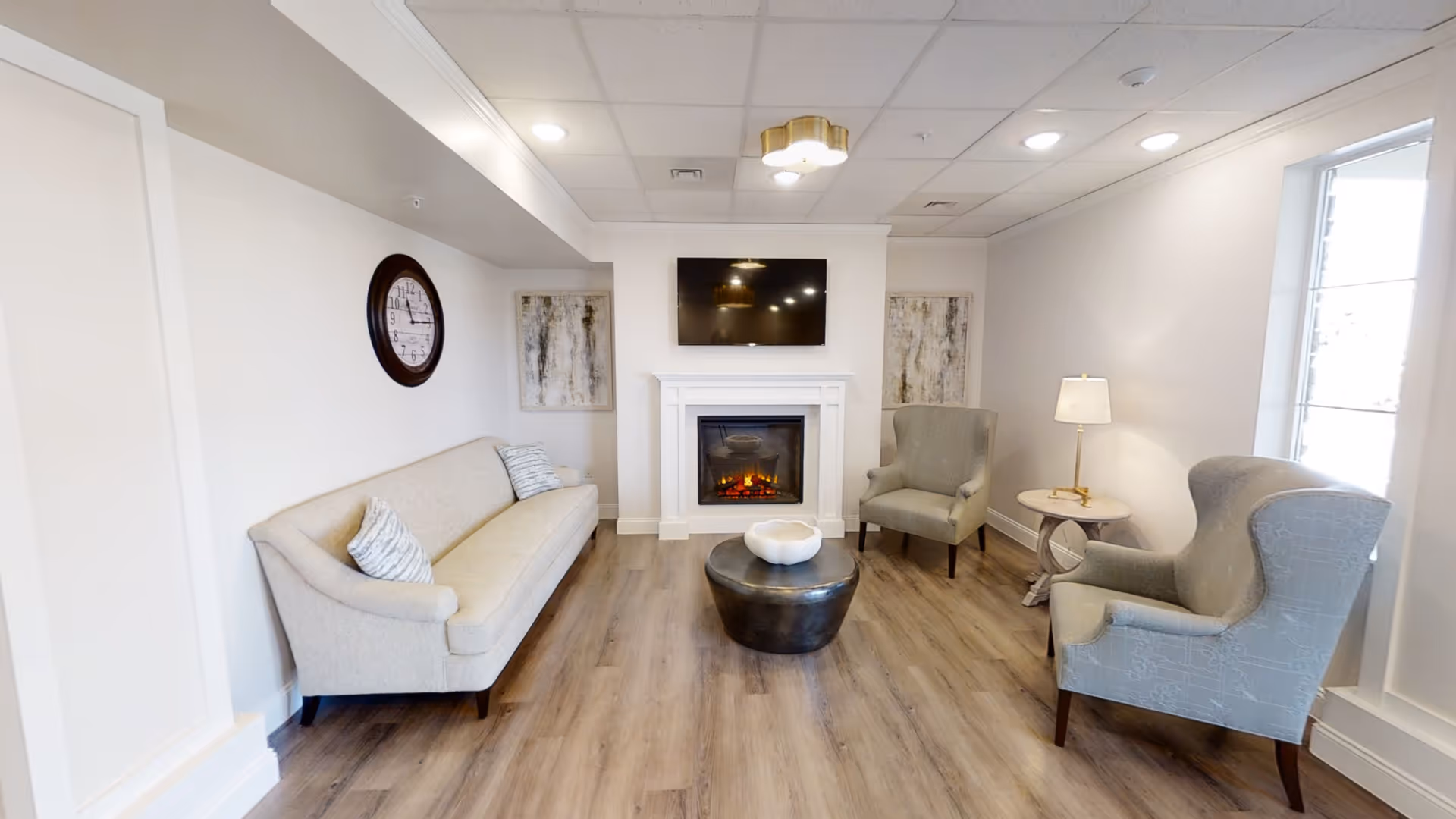 A cozy living room with a beige sofa on the left, two gray armchairs on the right, a round black coffee table in the center with a white decorative bowl, a white electric fireplace with a mounted flat-screen TV above it, two abstract paintings on either side of the fireplace, a round wall clock above the sofa, a small side table with a lamp next to one armchair, and a large window letting in natural light on the right.