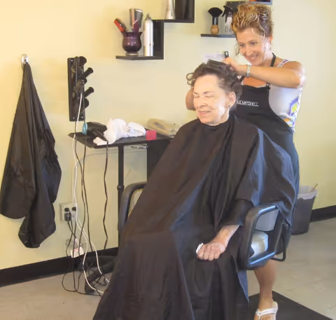 An elderly woman sitting in a salon chair with a black cape around her, getting her hair styled by a hairdresser in a salon setting with hair products and tools visible on shelves and a counter.