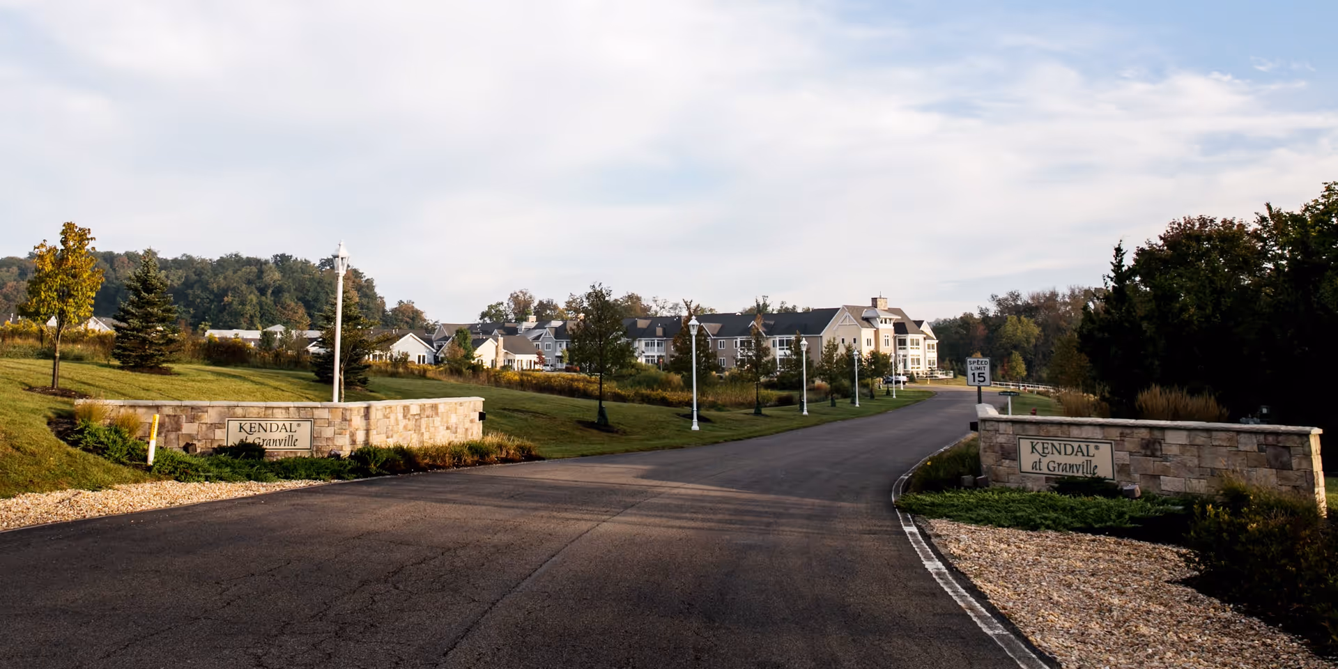 Entrance road leading to a senior living community with stone entrance walls and multi-story buildings in the distance.
