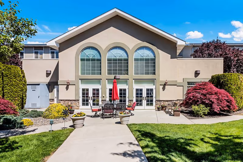 Exterior view of a senior living facility building with large arched windows and three sets of glass double doors. In front of the building is a concrete patio with outdoor furniture including a table, chairs, and a red umbrella. The surrounding area has well-maintained landscaping with green grass, bushes, and colorful plants under a clear blue sky.