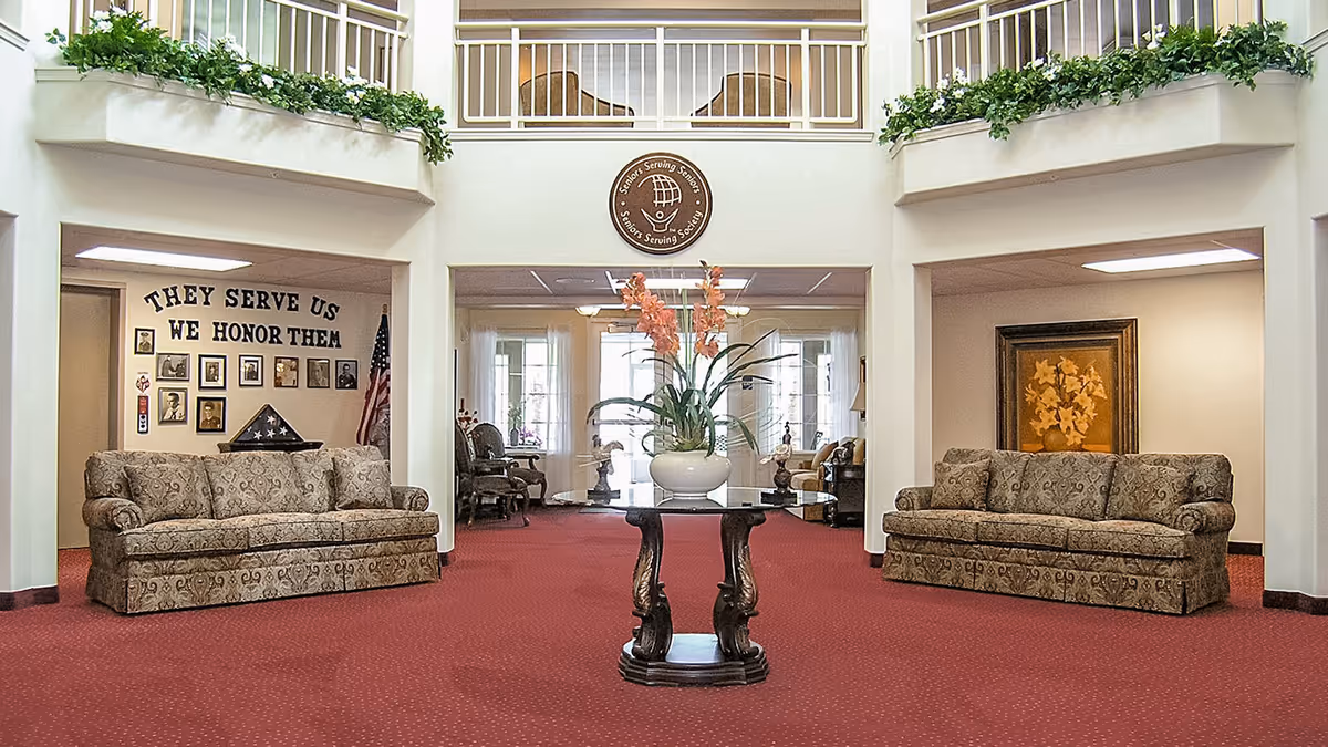 Spacious senior-living lobby with two patterned sofas flanking a central table holding a potted floral arrangement, red carpet, and an upper balcony.