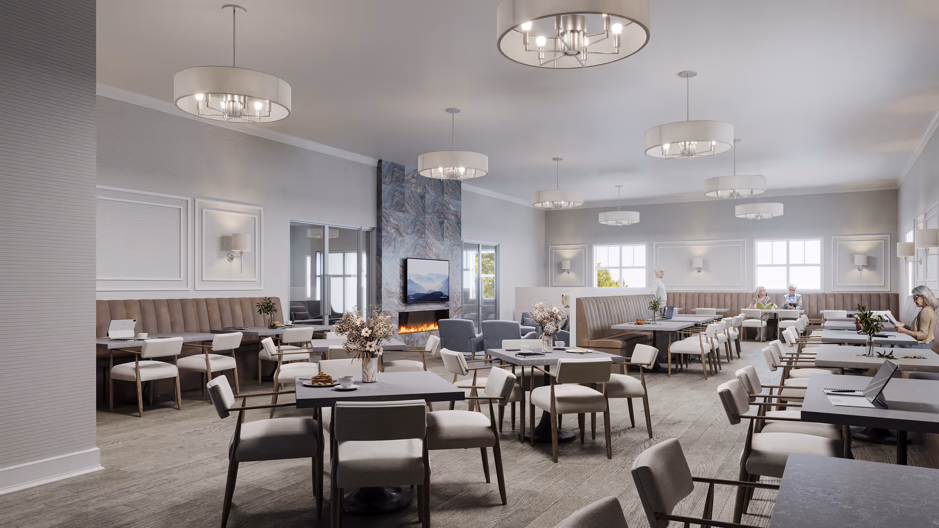 A spacious, modern dining room with multiple tables and chairs arranged neatly. The room features a large stone fireplace with a mounted flat-screen TV above it. There are several pendant lights hanging from the ceiling, and the walls are decorated with sconces and framed paneling. Large windows allow natural light to fill the space. A few elderly individuals are seated at tables, engaged in conversation or reading.