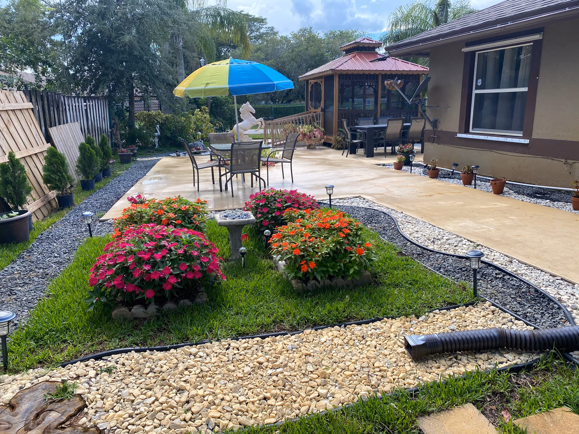 Outdoor patio area with a concrete walkway, colorful flower beds, and a table with chairs under a blue and yellow umbrella. There is a wooden gazebo with additional seating in the background, surrounded by greenery and potted plants.