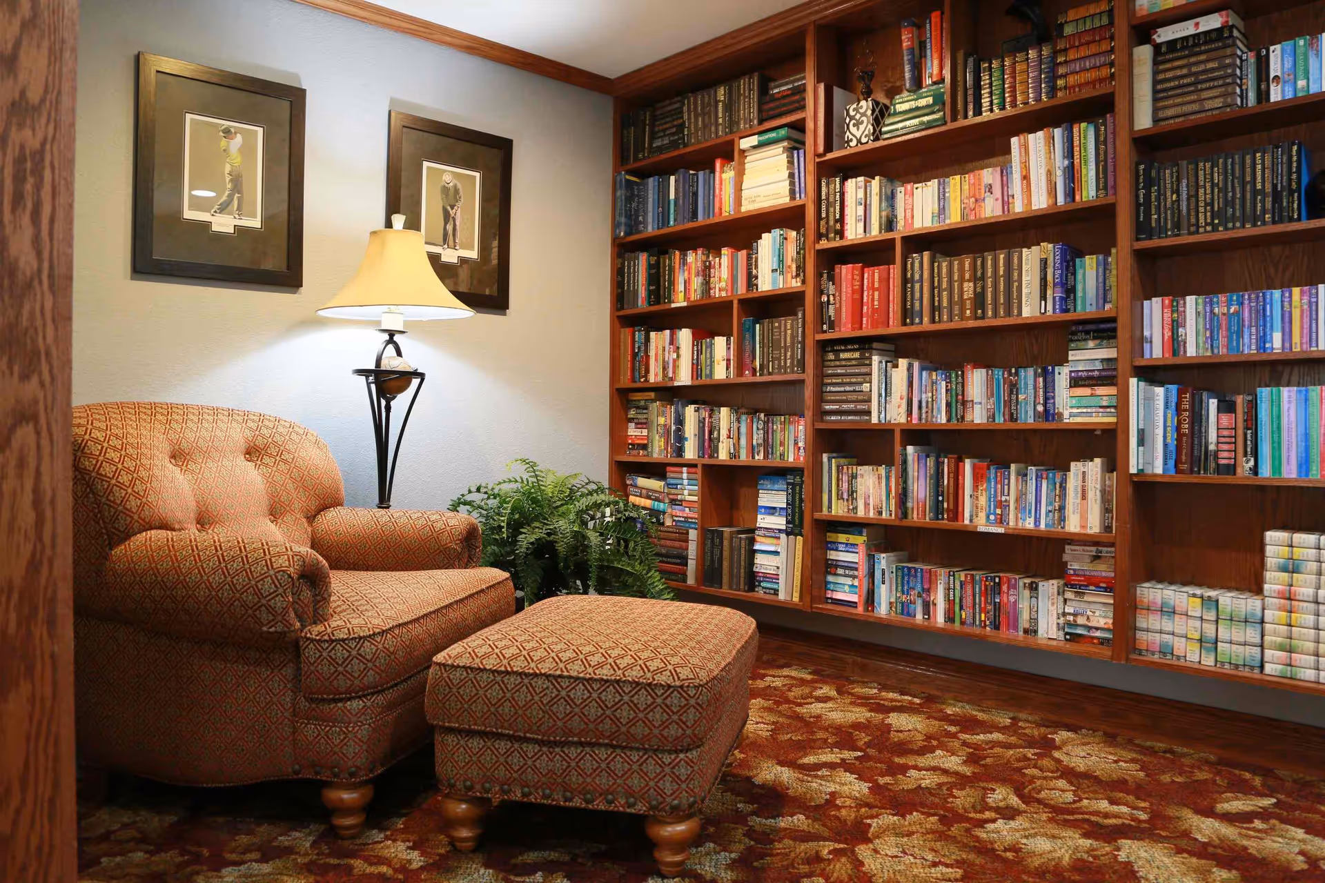 Cozy reading nook with an upholstered armchair and matching ottoman beside a floor lamp and framed pictures, facing built-in bookshelves filled with books.