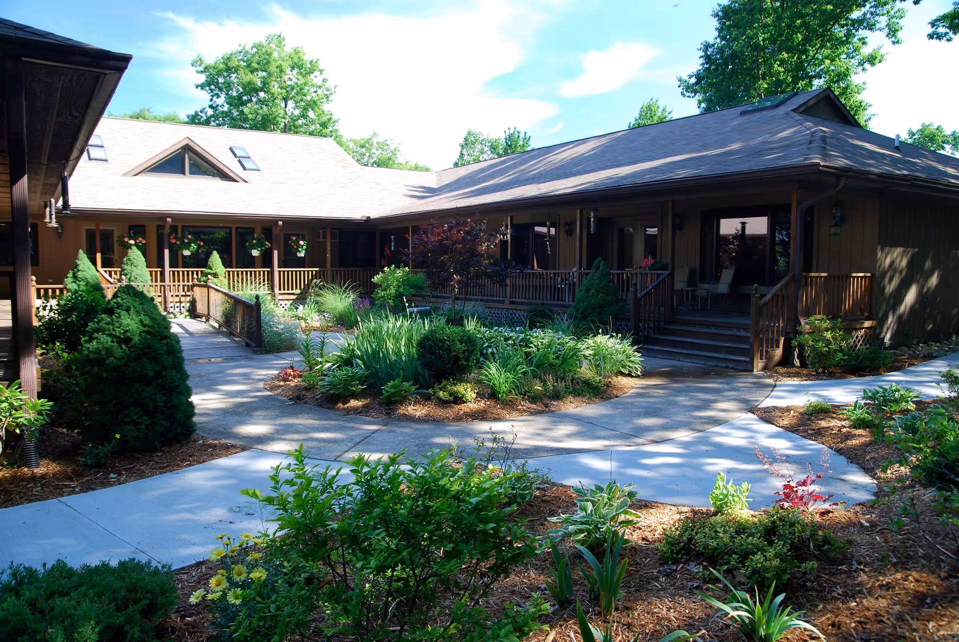 Sunlit courtyard with paved walkways, landscaped garden beds and a single-story wood-clad building with a covered porch.
