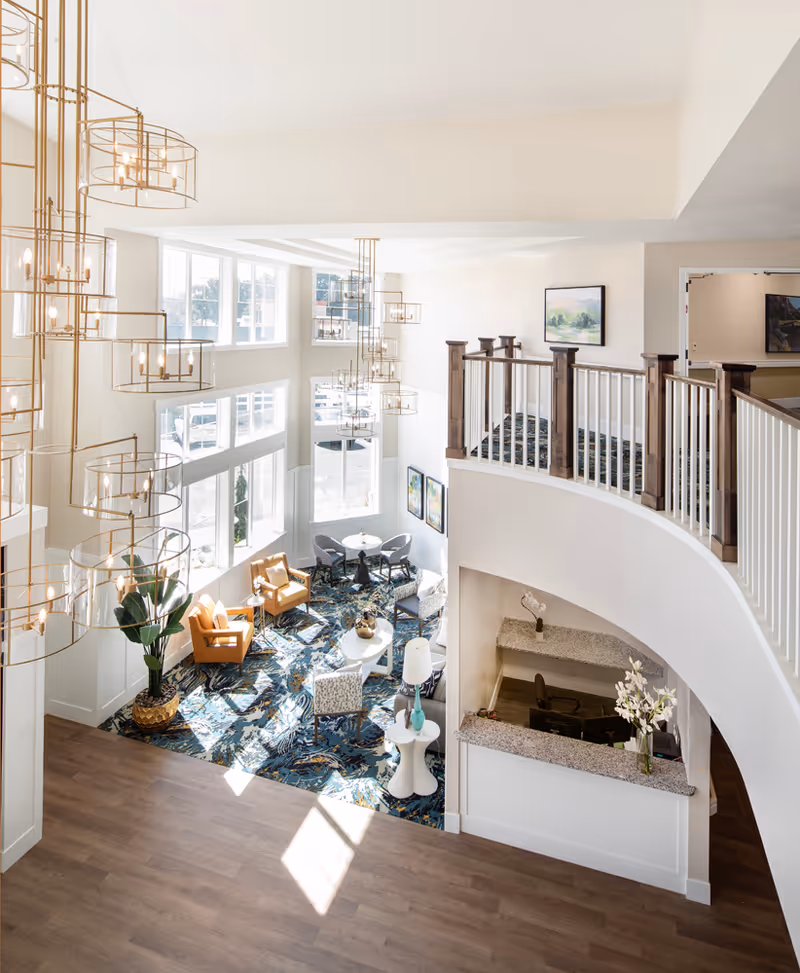 Bright two-story senior living lobby with large windows, multiple hanging chandeliers, a seating area on patterned carpet and a reception desk below a curved balcony.