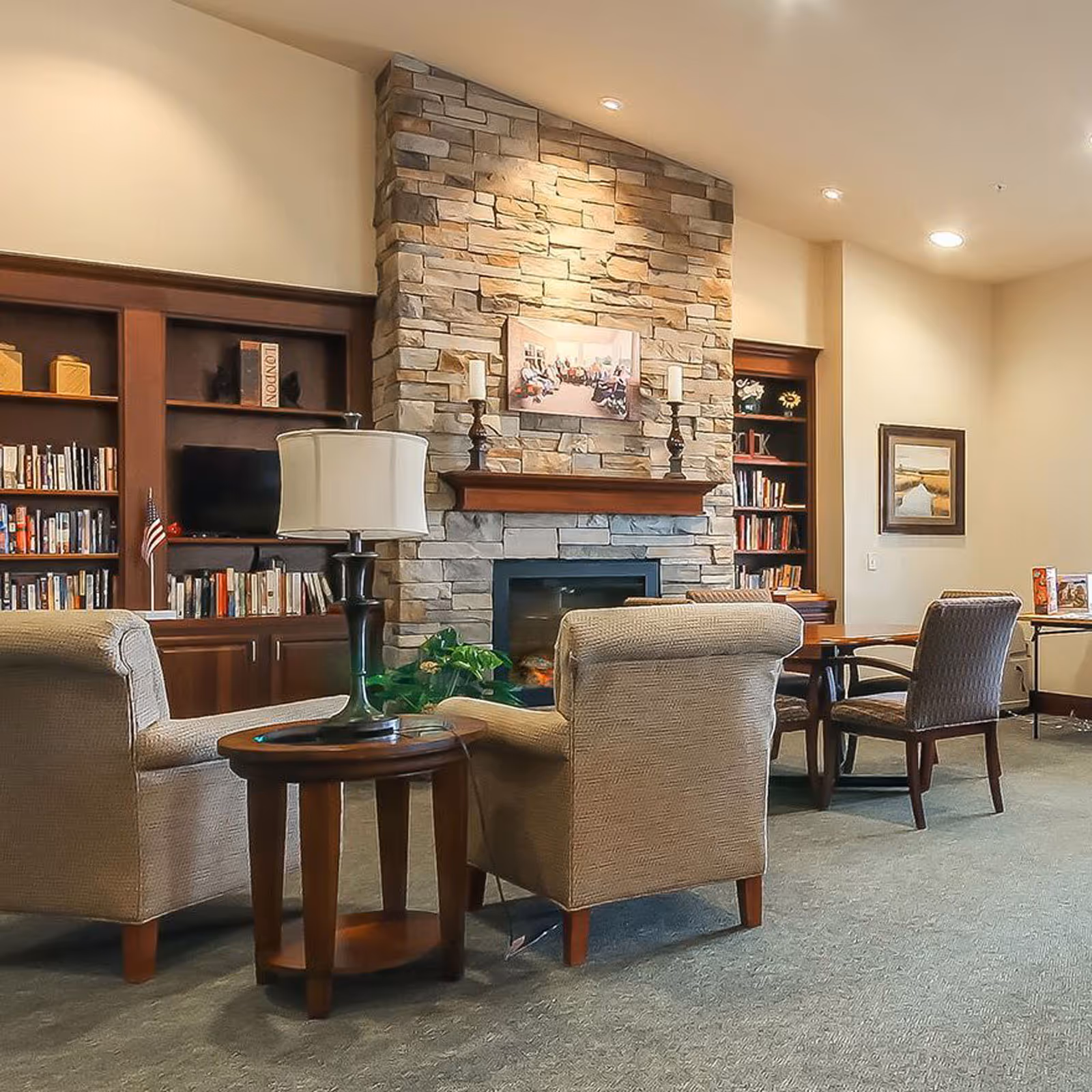 Cozy common living room with upholstered armchairs, a side table lamp, bookshelves and a stone fireplace.