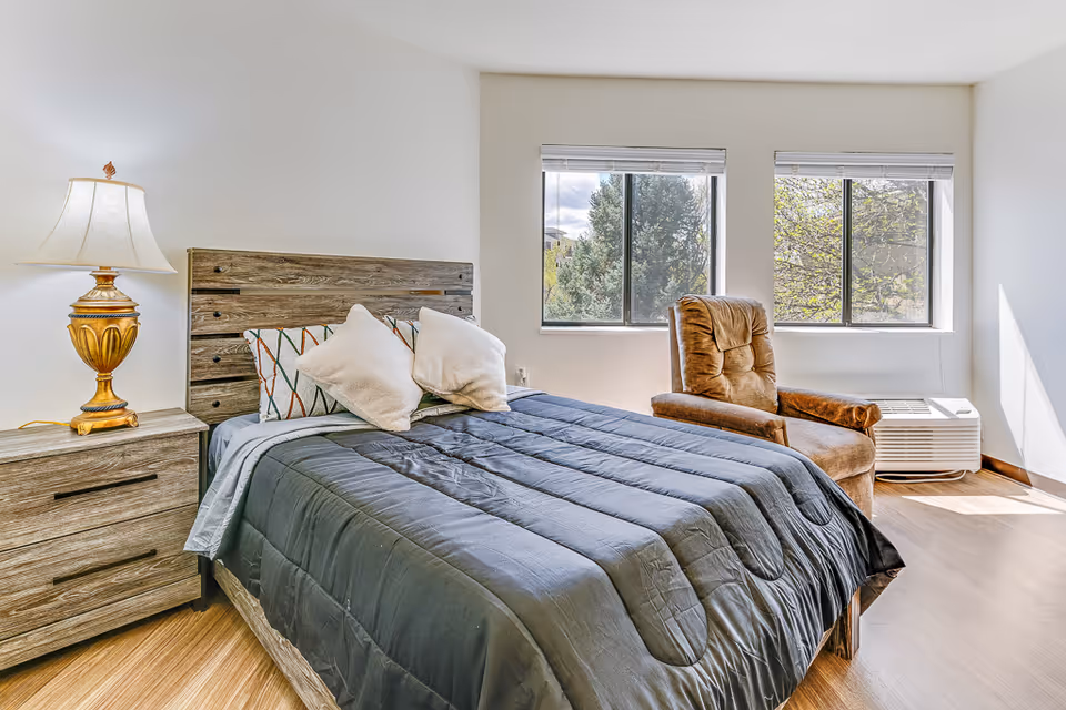 A bright, modern bedroom with a bed topped by a dark comforter and pillows, a wooden nightstand with a lamp, and a reclining armchair by two windows.