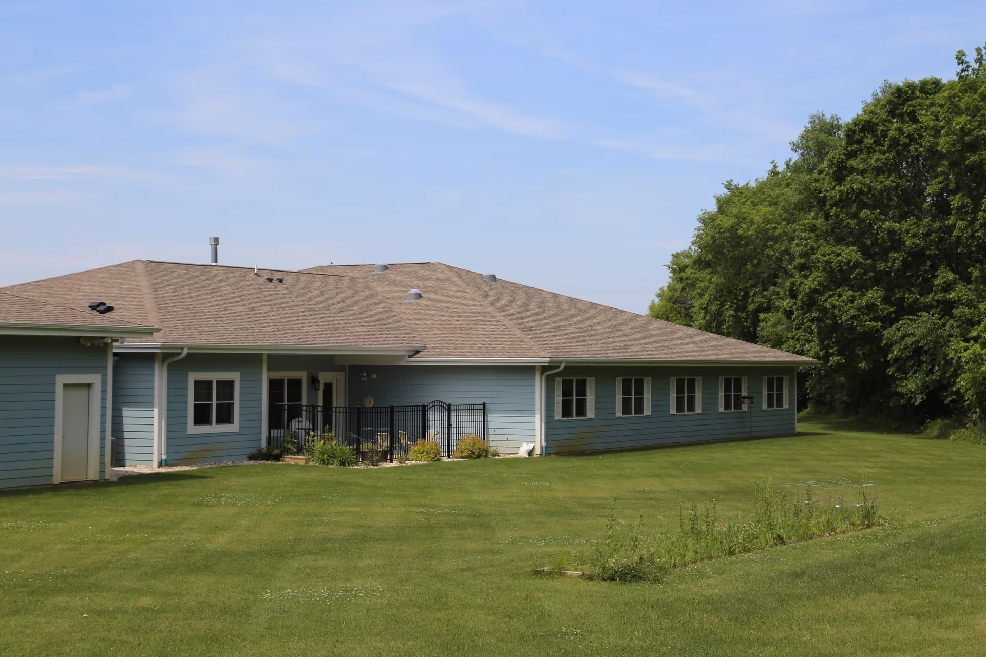 Exterior view of a single-story blue building with multiple windows and a brown shingled roof, surrounded by a large green lawn and trees under a clear blue sky.